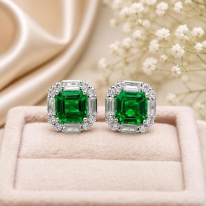 May include: A pair of stud earrings featuring a square-cut emerald green centre stone surrounded by a halo of baguette and round-cut clear white stones set in a polished silver-toned metal frame, creating a bright and reflective jewellery piece.
