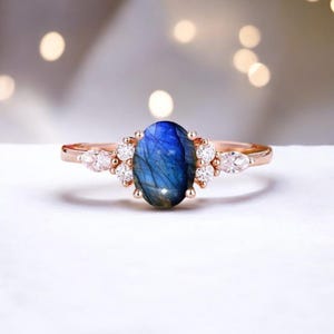 May include: Rose gold ring featuring an oval, blue labradorite gemstone. The ring is accented with small, round, and pear-shaped white gemstones. The ring's band is a delicate rose gold color, and the gemstones are set in a cluster design.