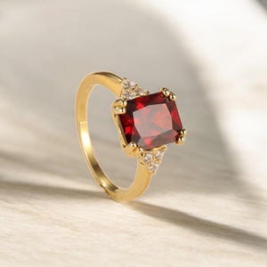 Może przedstawiać: Złoty pierścionek z dużym, prostokątnym, głęboko czerwonym kamieniem szlachetnym. Pierścionek ma małe, trójkątne skupiska mniejszych, przezroczystych kamieni po obu stronach głównego kamienia.