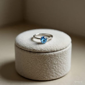 Może przedstawiać: Srebrny pierścionek z okrągłym, jasnoniebieskim kamieniem szlachetnym. Pierścionek leży na małym, okrągłym, kremowym aksamitnym pudełku. Obrączka pierścionka jest cienka i wypolerowana, a kamień szlachetny jest centralnym punktem biżuterii.