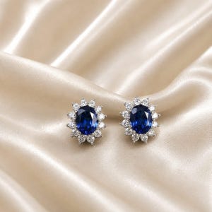 May include: A pair of stud earrings featuring a central oval-cut deep blue gemstone surrounded by a halo of small, clear, round-cut white stones. The earrings have a metallic setting that holds the stones in a classic floral-inspired arrangement.