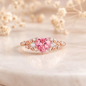 May include: A rose gold ring featuring a central heart-shaped pink gemstone flanked by two round clear stones. The band is designed with intricate leaf-like metalwork and small accent stones, creating a detailed botanical-inspired appearance.