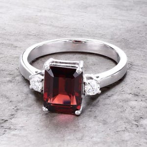 May include: A silver ring with a large, rectangular, deep red gemstone as the centerpiece. Two smaller, round, clear gemstones flank the main stone. The ring is set on a gray surface.