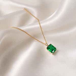 May include: A gold chain necklace featuring a rectangular emerald-cut green gemstone pendant. The pendant is held by four small prongs in a gold setting, suspended from a delicate gold link chain that rests against a smooth, draped fabric.