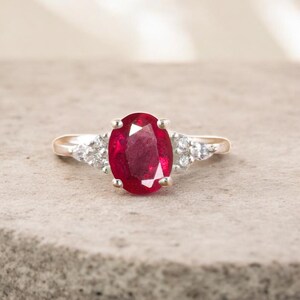 Może przedstawiać: Złoty pierścionek z dużym, owalnym rubinem w centrum. Po obu stronach rubinu znajdują się trójkątne skupiska mniejszych, białych kamieni. Projekt pierścionka jest umieszczony na neutralnym tle, podkreślając bogaty czerwony odcień rubinu.