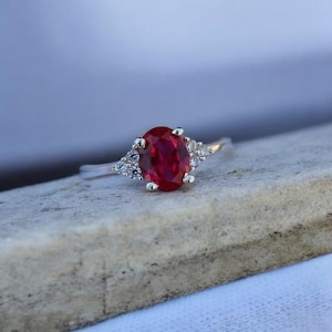 May include: A silver ring with a large oval, deep red gemstone as the focal point. The ring is flanked by smaller, clear gemstones on each side. The ring is set on a light-colored, textured surface.