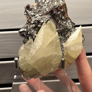 Puede incluir: Un espécimen mineral que presenta un grupo de cristales. Los cristales son de color amarillo pálido y tienen bordes afilados y puntiagudos. El espécimen está montado sobre un soporte de acrílico transparente, mostrando la belleza natural de la formación geológica.
