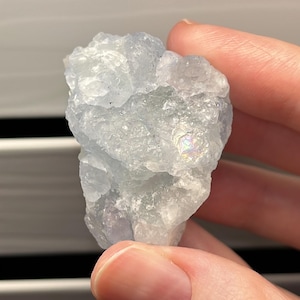 May include: A light blue, raw fluorite crystal with a rough, textured surface. The crystal has areas of iridescence, displaying a rainbow effect. The mineral is held in a hand, showcasing its natural form and color variations.