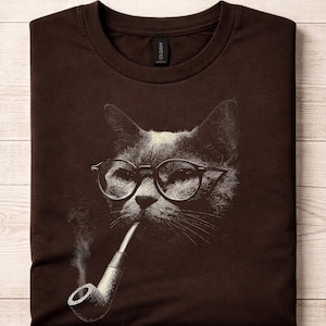 May include: Brown t-shirt featuring a detailed illustration of a cat wearing glasses and smoking a pipe. The cat's face is in black and white, with the glasses and pipe in a lighter shade. The shirt is folded on a light wood surface.