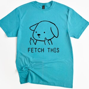 May include: A turquoise t-shirt with a cartoon dog illustration giving the middle finger. The black graphic includes the text "FETCH THIS". The shirt is made of a soft material. The design is centered on the front.