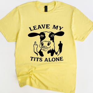 May include: A yellow t-shirt featuring a black and white cow graphic giving the middle finger. The text "LEAVE MY TITS ALONE" is printed above and below the cow illustration. The shirt has a crew neck and short sleeves.
