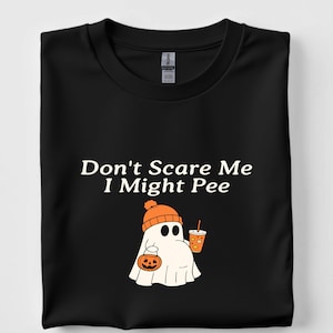 May include: Black t-shirt with the text "Don't Scare Me I Might Pee." The shirt features a cartoon ghost wearing an orange beanie and holding a pumpkin and a drink. The ghost has black eyes and a white body.
