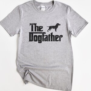 May include: A light gray t-shirt with the text "The Dogfather" in a distressed black font, accompanied by a black silhouette of a dog. The shirt has a crew neck and short sleeves.