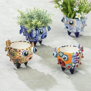 May include: Four ceramic planters in the shape of fish, each with a unique design and color scheme. The planters have large, expressive eyes and are supported by three small legs. Two planters contain greenery and flowers.