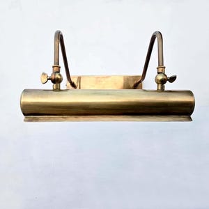 May include: A vintage brass picture light fixture. The light has a cylindrical body with two curved arms and adjustable knobs. The fixture is mounted on a rectangular backplate and is designed to illuminate artwork or other displays.