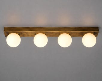 Puede incluir: Una lámpara lineal con una base rectangular de metal en tono latón envejecido y cuatro globos esféricos de vidrio blanco espaciados uniformemente. Los globos emiten una luz suave y cálida, diseñada para su montaje en posición horizontal.