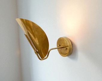 May include: A brass wall sconce with a curved, leaf-shaped shade and a circular base. The sconce is mounted on a white wall, and the light is on, casting a warm glow. The design is mid-century modern.