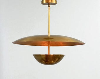 May include: A brass pendant light fixture with a large, shallow, saucer-shaped shade and a smaller, bowl-shaped element below. The fixture has a gold finish and a central rod for hanging. The design is modern and minimalist.