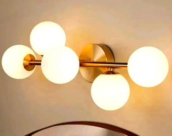 May include: A modern wall-mounted light fixture with five spherical, frosted glass shades. The fixture has a gold-toned metal finish, with a circular base and horizontal bars supporting the globes. The light is designed for interior use.
