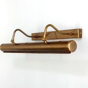 May include: A vintage-style picture light fixture with a bronze finish. The fixture has a rectangular backplate and a cylindrical light housing. Two curved arms extend from the backplate to support the light housing. The light is designed to illuminate artwork.