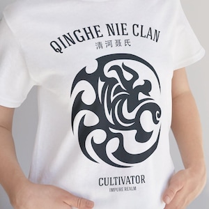 Könnte beinhalten: Weißes T-Shirt mit einem schwarzen Grafikdesign. Das Design zeigt den Text "QINGHE NIE CLAN" und "CULTIVATOR" sowie ein kreisförmiges Emblem mit stilisierten Formen und chinesischen Schriftzeichen. Das T-Shirt ist aus weichem Material.