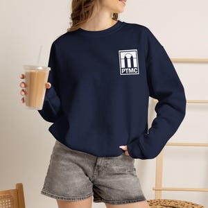 May include: A navy blue long-sleeve sweatshirt featuring a white rectangular graphic on the upper left chest. The graphic displays an architectural arch design above the text PTMC. The sweatshirt is paired with gray denim shorts.