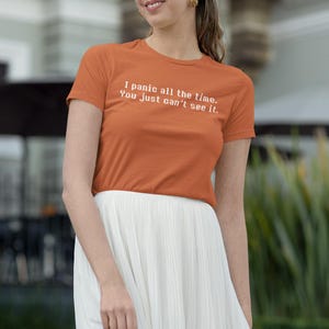 May include: An orange t-shirt with the text "I panic all the time. You just can't see it." is worn with a white pleated skirt. The person is wearing sunglasses and gold earrings.