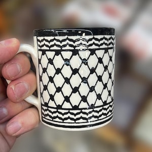 May include: A small, white ceramic mug with a black geometric pattern. The mug has a handle and a black rim. The design features a repeating pattern of diamonds and curved lines. The mug is held in a hand.