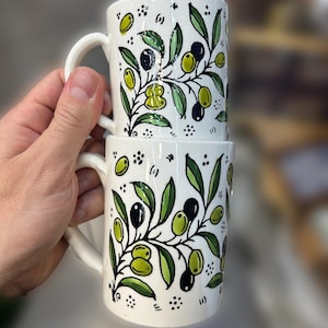 May include: Two white ceramic mugs stacked, each adorned with a hand-painted olive branch design. The design features green and black olives, green leaves, and delicate black stems. The mugs have a curved handle.