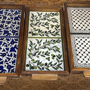 Puede incluir: Tres bandejas de madera con inserciones de baldosas de cerámica decorativas. Las baldosas presentan diseños florales, de ramas de olivo y geométricos en azul, verde, negro y blanco. Cada bandeja tiene forma rectangular con marco y asas de madera.