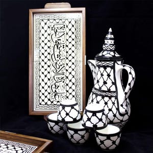 Può includere: Un set da caffè in ceramica bianca e nera con una caffettiera alta e ornata, piccole tazze e un vassoio decorativo. Il set presenta un motivo geometrico e calligrafia araba. Il vassoio e le tazze sono disposti davanti a una piastrella incorniciata con più calligrafia.