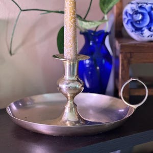 May include: A vintage brass candlestick holder with a circular base and a handle. A tall, yellow candle with white speckles sits in the holder. The background includes a blue vase and a decorative ceramic piece.
