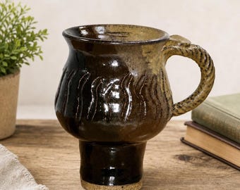 May include: A ceramic mug with a bulbous body and a narrow base, featuring a glossy dark brown and tan glaze. The surface has vertical textured indentations around the middle, and the handle has a distinct twisted rope-like appearance.