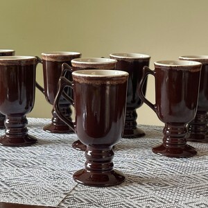 May include: A set of seven dark brown ceramic mugs with a pedestal base and a handle. Each mug features a cylindrical body and a distinct, textured, light-colored rim detail. The mugs are arranged in a group, showcasing their glossy finish.
