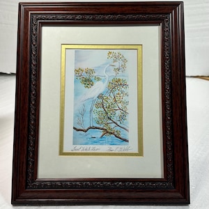 May include: A framed watercolour painting of a Great White Heron. The heron is depicted perched on a tree branch, set against a blue water and sky backdrop. The artwork is presented in a dark brown, ornate wooden frame.