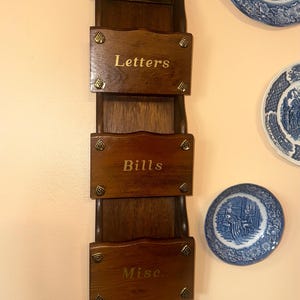 May include: A dark wooden wall-mounted organizer with three labeled compartments: "Letters", "Bills", and "Misc.". The organizer has brass accents and a decorative top. Blue and white decorative plates are visible in the background.