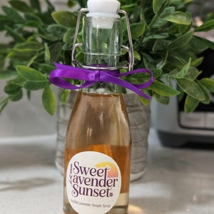 Puede incluir: Una pequeña botella de vidrio llena de un líquido ámbar claro, etiquetada como "Sweet Lavender Sunset Vanilla Lavender Simple Syrup". La botella tiene un tapón blanco, un cierre metálico y una cinta morada. Fondo de follaje verde.