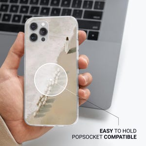 May include: Clear phone case featuring a serene beach scene with a person in white walking along the shore, followed by a flock of sheep. The case is labeled "EASY TO HOLD POPSOCKET COMPATIBLE".