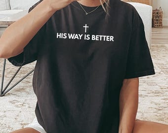 May include: Black t-shirt with the text "HIS WAY IS BETTER" and a cross symbol in white. The person is wearing white socks with black stripes. The person is sitting on a patterned rug.