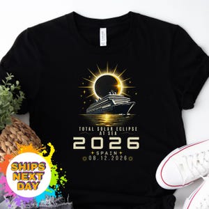 May include: A black t-shirt featuring a graphic of a cruise ship sailing under a total solar eclipse. The text reads TOTAL SOLAR ECLIPSE AT SEA 2026 SPAIN 08.12.2026. The design includes a glowing sun, stars, and golden light reflecting on the water.