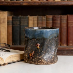 May include: A handcrafted ceramic mug with a dark blue glazed rim and a textured, brown body. The mug is adorned with small, amber-coloured stones. It's placed in front of a backdrop of antique books, creating a cosy, literary atmosphere.