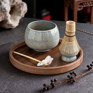 Può includere: Un set da tè matcha su un vassoio di legno. Il set include una ciotola in ceramica blu e bianca, un frullino di bambù, un cucchiaio di bambù e un poggia cucchiaio in ceramica bianca. Il vassoio è rotondo e in legno scuro.