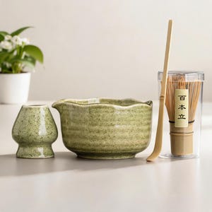 May include: A matcha tea set comprising a green ceramic bowl, a whisk, a scoop, and a small ceramic holder. The whisk is in a clear container. The set is for preparing matcha tea.