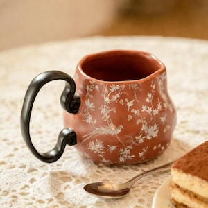 May include: A terracotta-colored ceramic mug with a black handle. The mug is decorated with a white floral and bird pattern. A silver spoon and a piece of tiramisu are in the foreground, suggesting a tea or coffee setting.