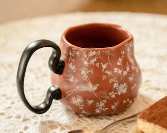 May include: A terracotta ceramic mug with a black handle, decorated with a white floral and bird pattern. A silver spoon and tiramisu are in the foreground. The scene suggests a tea or coffee break.