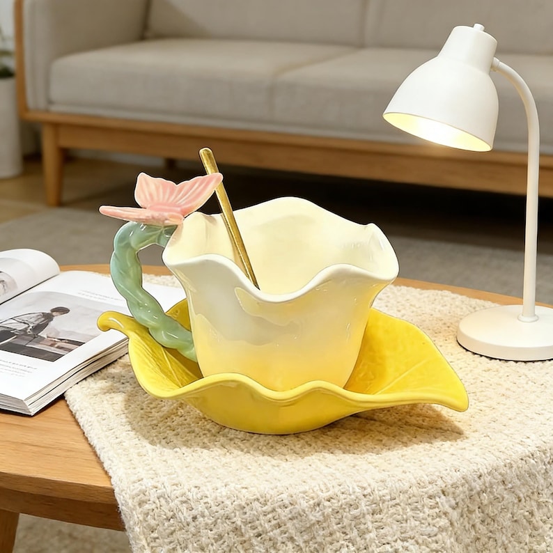 May include: A yellow ceramic teacup and saucer set with a floral design. The cup has a petal-like shape and a butterfly handle. A gold spoon rests inside the cup. The saucer is leaf-shaped.