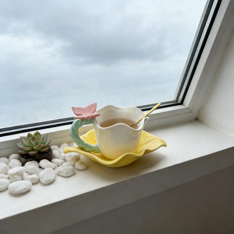 May include: A white teacup with a flower-shaped rim and a butterfly handle, on a yellow saucer. A gold spoon rests in the cup, filled with tea. A small succulent plant and white stones are in the background.