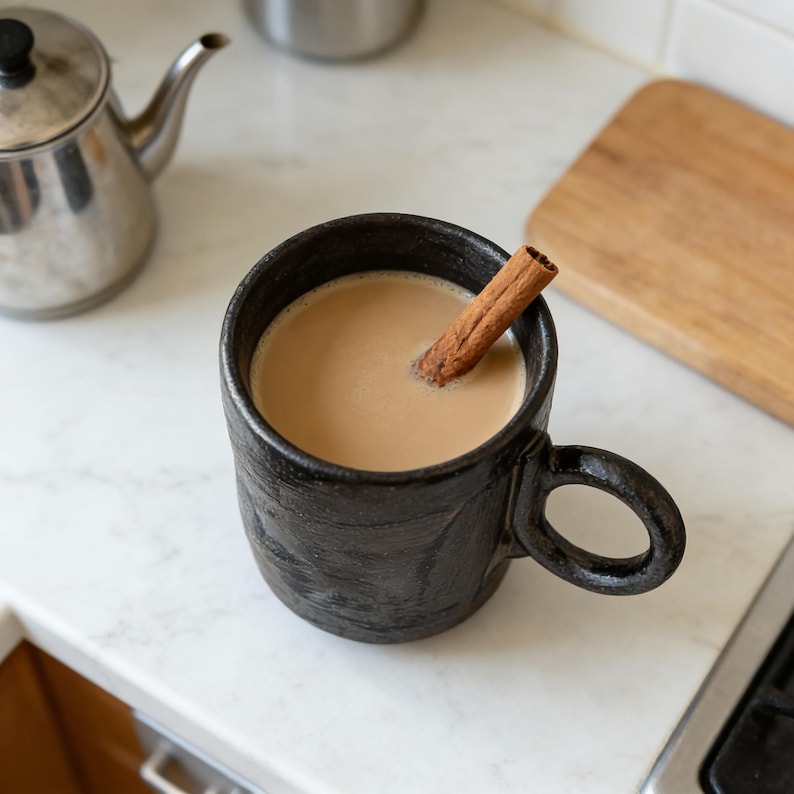 Op de afbeelding: Een zwarte keramische mok gevuld met een romig drankje en een kaneelstokje. De mok heeft een textuur en een grote handgreep. Een roestvrijstalen theepot en een houten snijplank staan op de achtergrond.