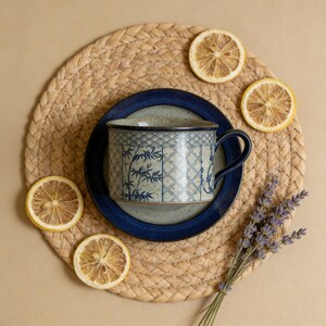 May include: A ceramic mug with a blue bamboo design sits on a matching saucer. The mug and saucer are on a woven placemat, surrounded by dried lemon slices and a sprig of lavender. The mug has a dark blue handle and rim.