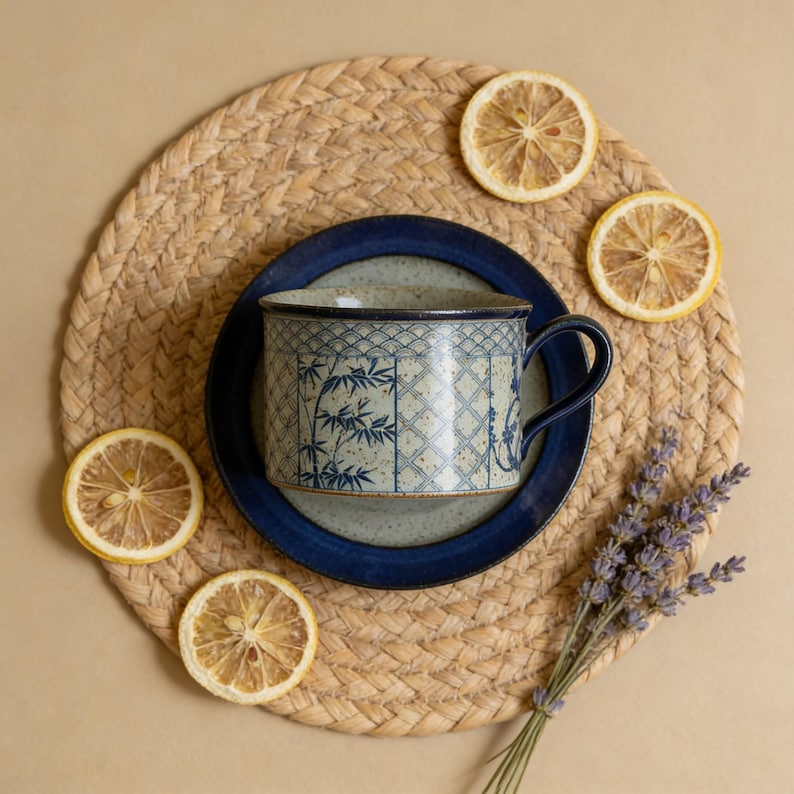 May include: A ceramic mug with a blue bamboo design sits on a matching saucer. The mug and saucer are on a woven placemat, surrounded by dried lemon slices and a sprig of lavender. The mug has a dark blue handle and rim.